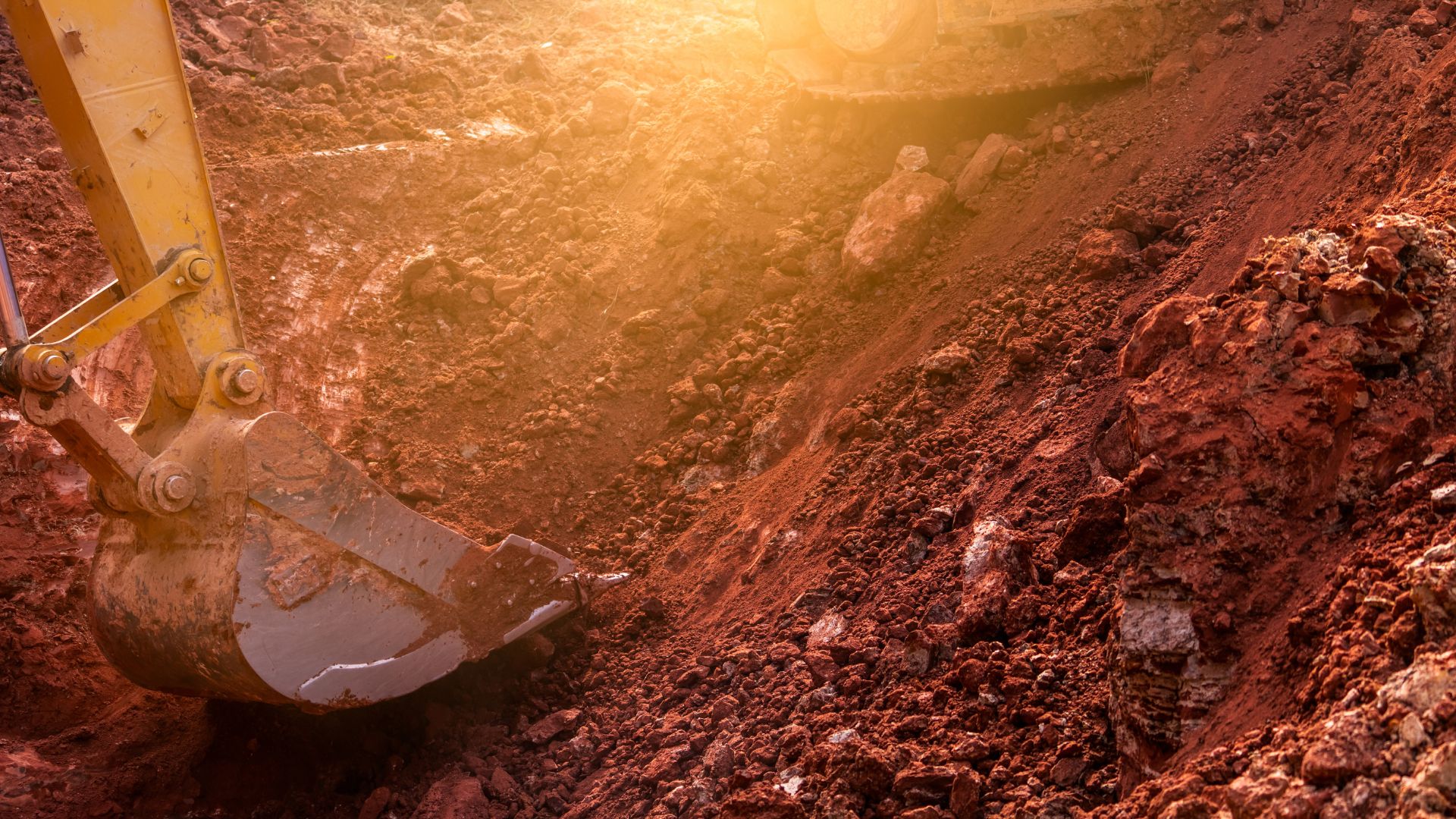 Excavator bucket digging into red rocky soil with warm sunlight